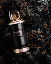 Black perfume extrait bottle with gold cap, gold logo label, and textured base ring
